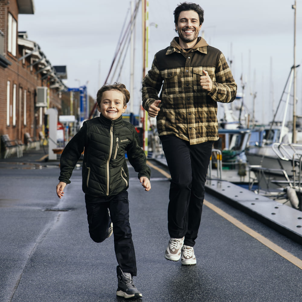 Zwei Personen in grüner Outdoorbekleidung beim gemeinsamen Laufen an einem Hafen, funktionale Freizeit- und Jagdbekleidung für Erwachsene und Kinder.