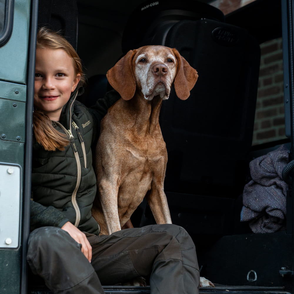 Kind in grüner Jagdbekleidung mit Jagdhund im Geländewagen, passend für Jagd, Revier und Outdoor-Aktivitäten.